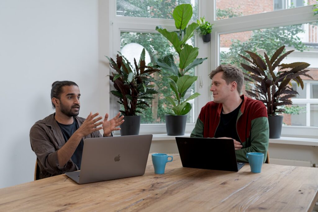 Two men collaborate at a table, each using a laptop, discussing proposal automation and teamwork strategies.